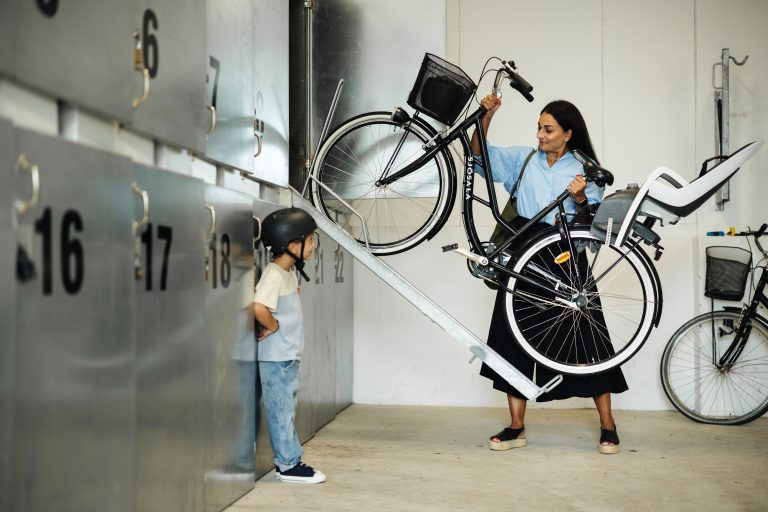Kvinna lägger in sin cykel i ett cykelskåp. Bredvid henne står ett barn i cykelhjälm och kollar.