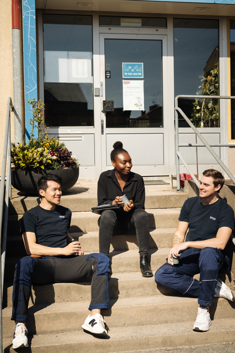 En grupp människor iklädda Bostadsbolagets kläder som sitter på trappan till en byggnad och ler.