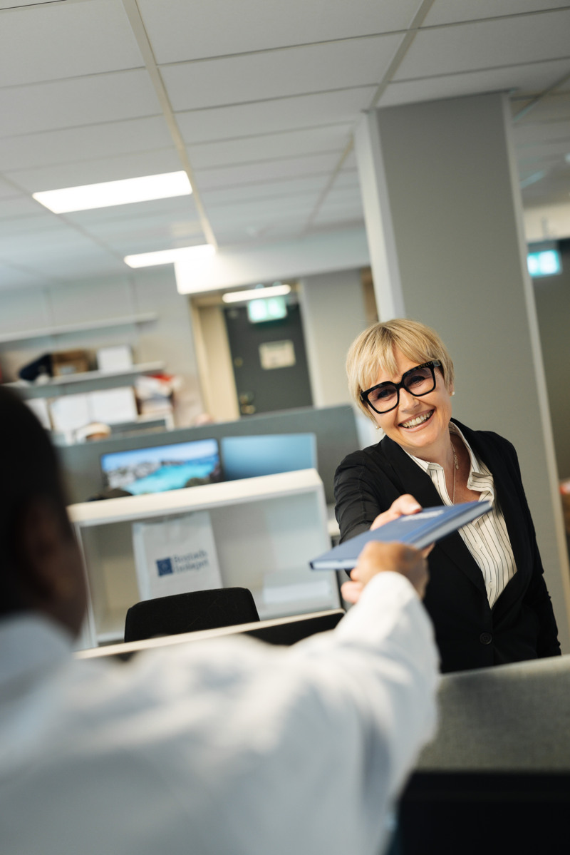 En leende kvinna i kostym räcker över ett anteckningsblock till en man. 