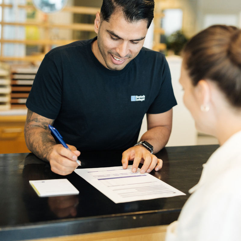 En leende man står vid ett skrivbord och skriver under ett papper. Framför honom står en leende kvinna. 