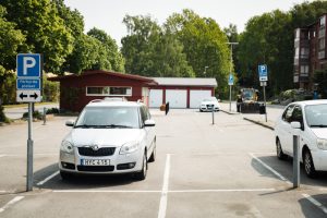 Två vita bilar som står parkerade på en parkeringsplats. I bakgrunden syns en parkeringsskylt, ett miljörum och gröna träd.