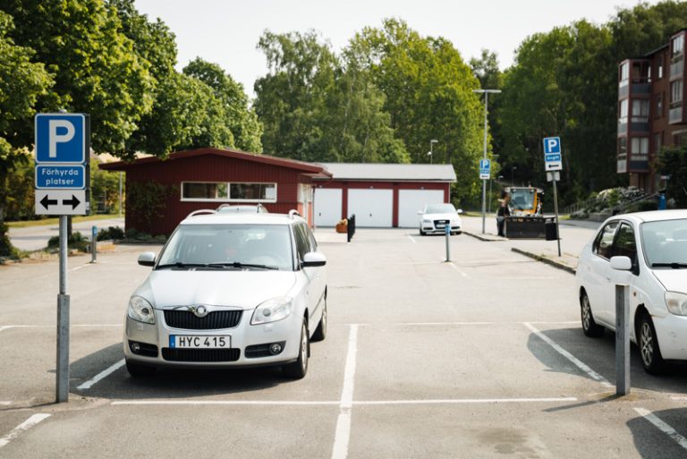 Två vita bilar som står parkerade på en parkeringsplats. I bakgrunden syns en parkeringsskylt, ett miljörum och gröna träd.