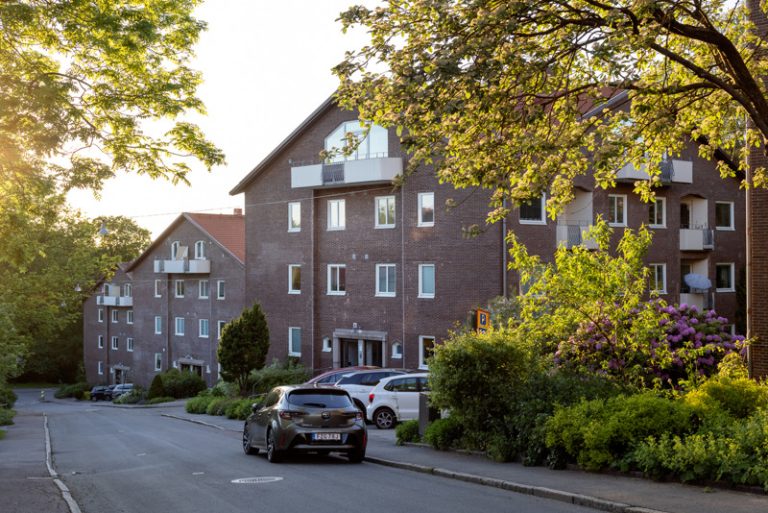 Tre bruna hus på rad i en nedförsbacke. Grönskande träd längs med sidorna och en bild som står parkerad på vägen.