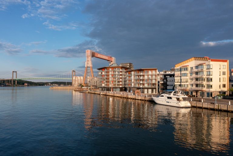 Hus längs med kajen. I havet finns en båt och i bakgrunden syns en kran och en båt.