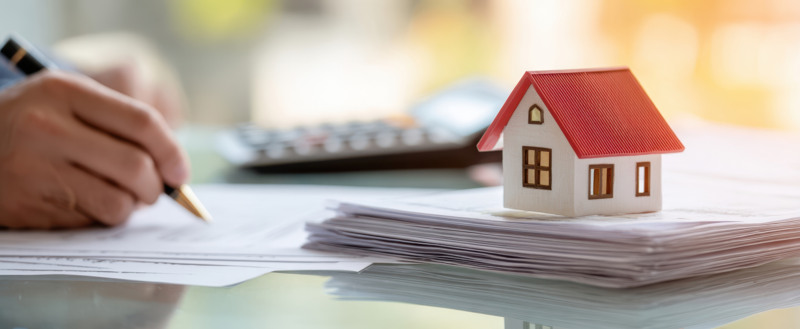 Bild på ett par händer som skriver på ett papper. I förgrunden syns en hög med papper, en miniräknare och en figurin av ett hus. 
