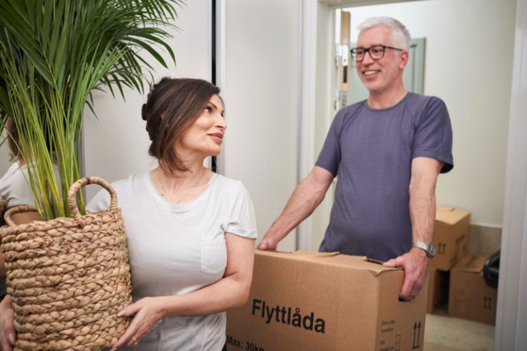 En leende man och kvinna som håller i en växt respektive flyttkartong. I bakgrunden syns ännu fler flyttkartonger. 