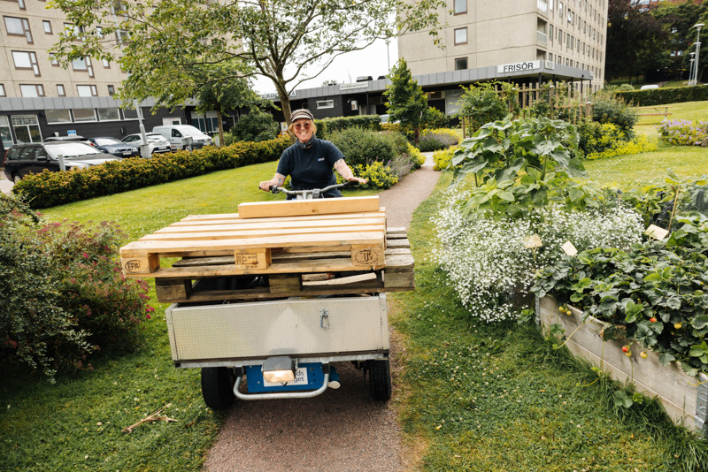 Bostadsbolagets miljövärd Pia sitter på en flakmoped och ler mot kameran. I bakgrunden syns pallkragar, gröna växter och beiga fastigheter. 