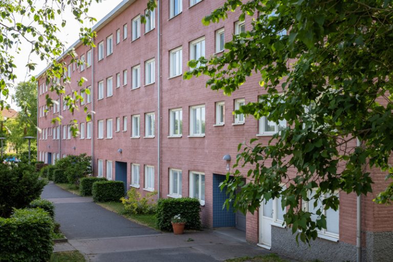 En byggnad i ljust tegel med många fönster och buskar.