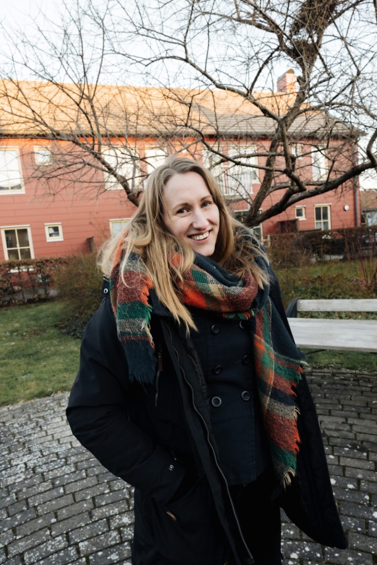 Sara, Bostadsbolagets säkerhetschef, tittar in i kameran och ler. Bakom henne syns ett rött hus, en bänk och ett stort träd. 