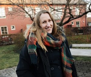 Sara, Bostadsbolagets säkerhetschef, tittar in i kameran och ler. Bakom henne syns ett rött hus, en bänk och ett stort träd. 