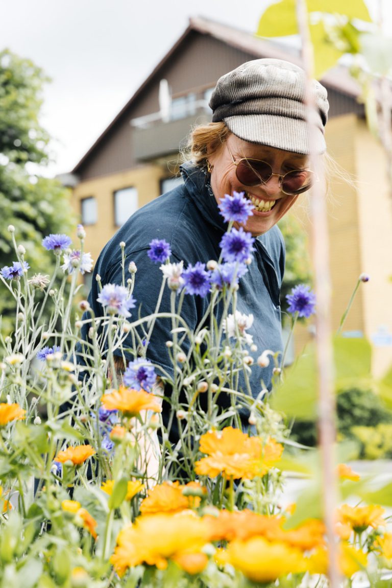 Bostadsbolagets miljövärd Pia står och ler framför ett par blommor i lila och gula nyanser. Bakom henne syns ett hus i gult och brunt tegel. 