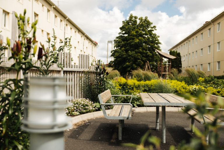 Solig sommardag i Bostadsbolagets bostadskvarter i Rambergsstaden. På bilden syns en sittgrupp, gröna växter och gula lägenhetsbygnader. 