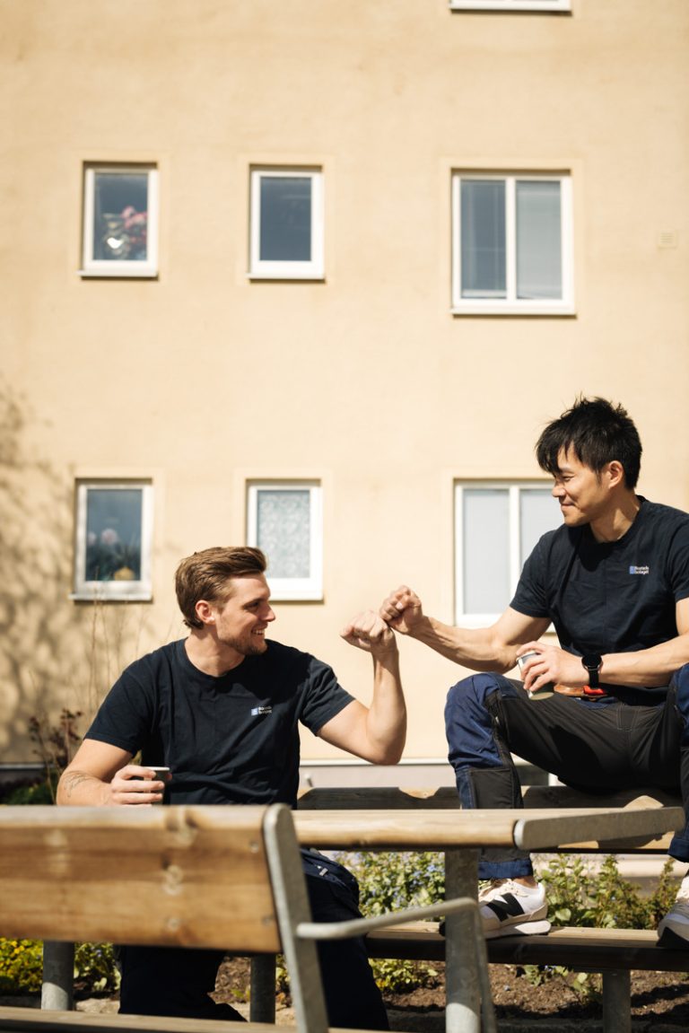 Två män sitter på en bänk framför en gul lägenhetsbyggnad och ler mot varandra.