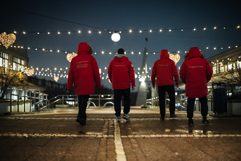 Bostadsbolagets trygghetsvärdar i Biskopsgården, iklädda röda jackor där det står "Trygghetsvärd" på ryggen. I bakgrunden syns juliga ljusdekorationer.