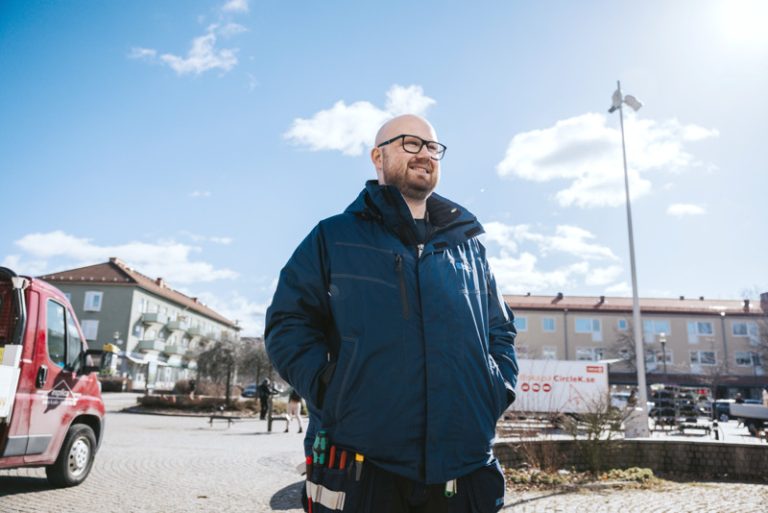 Bostadsbolagets fastighetsvärd Daniel på torget i Kyrkbyn. Daniel har på sig Bostadsbolagets arbetskläder och ser glad ut.