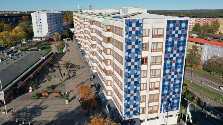 En flygbild av Skivhuset i Biskopsgården, som är ett högt lägenhetshus med färgglada solpaneler.