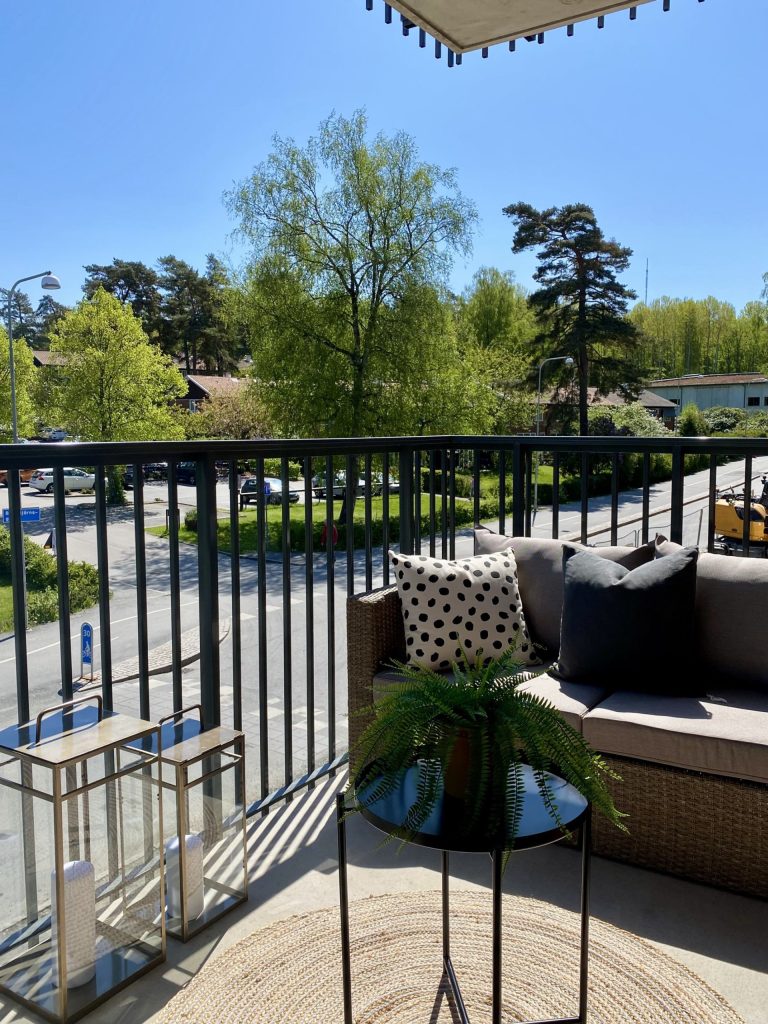 En balkong med en soffa och ett bord på den. I bakgrunden syns en blå himmel, en tom gata och ett par gröna träd.