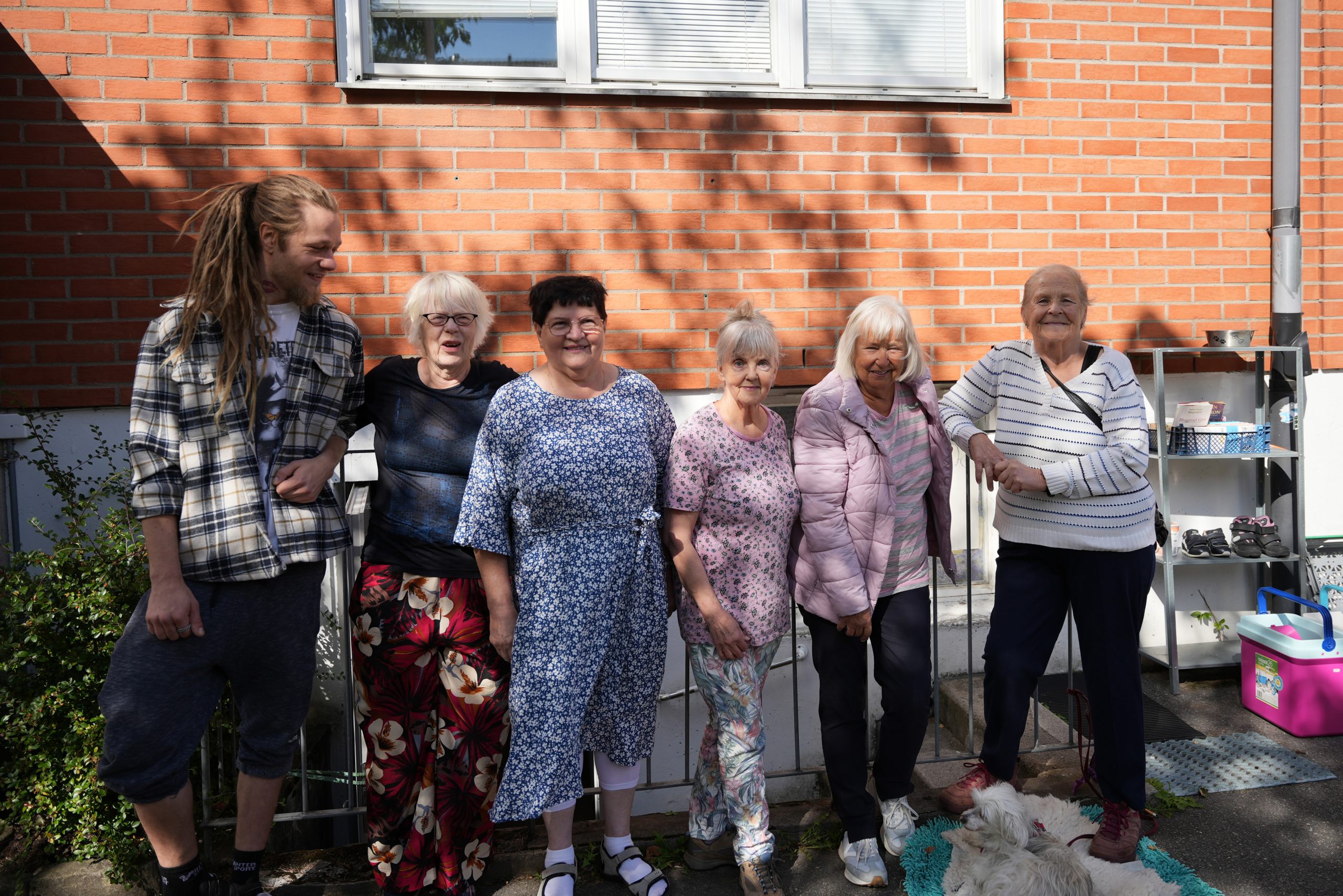 En grupp äldre kvinnor och en ung man till vänster som står bredvid varandra och ler framför ett loppisbord. 
