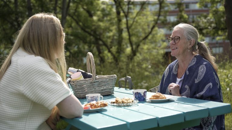 Två kvinnor sitter vid ett bord med tallrikar med fika och ler mot varandra. I bakgrunden syns gröna träd.