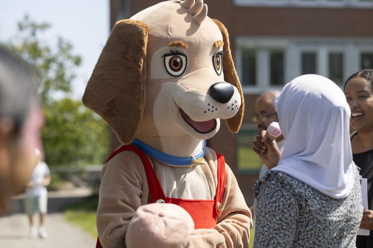 Bostadsbolagets maskot Trygga, en hund med röda hängselbyxor och brun päls. I den suddigare bakgrunden syns ett par skrattande personer och en lägenhetsbyggnad. 