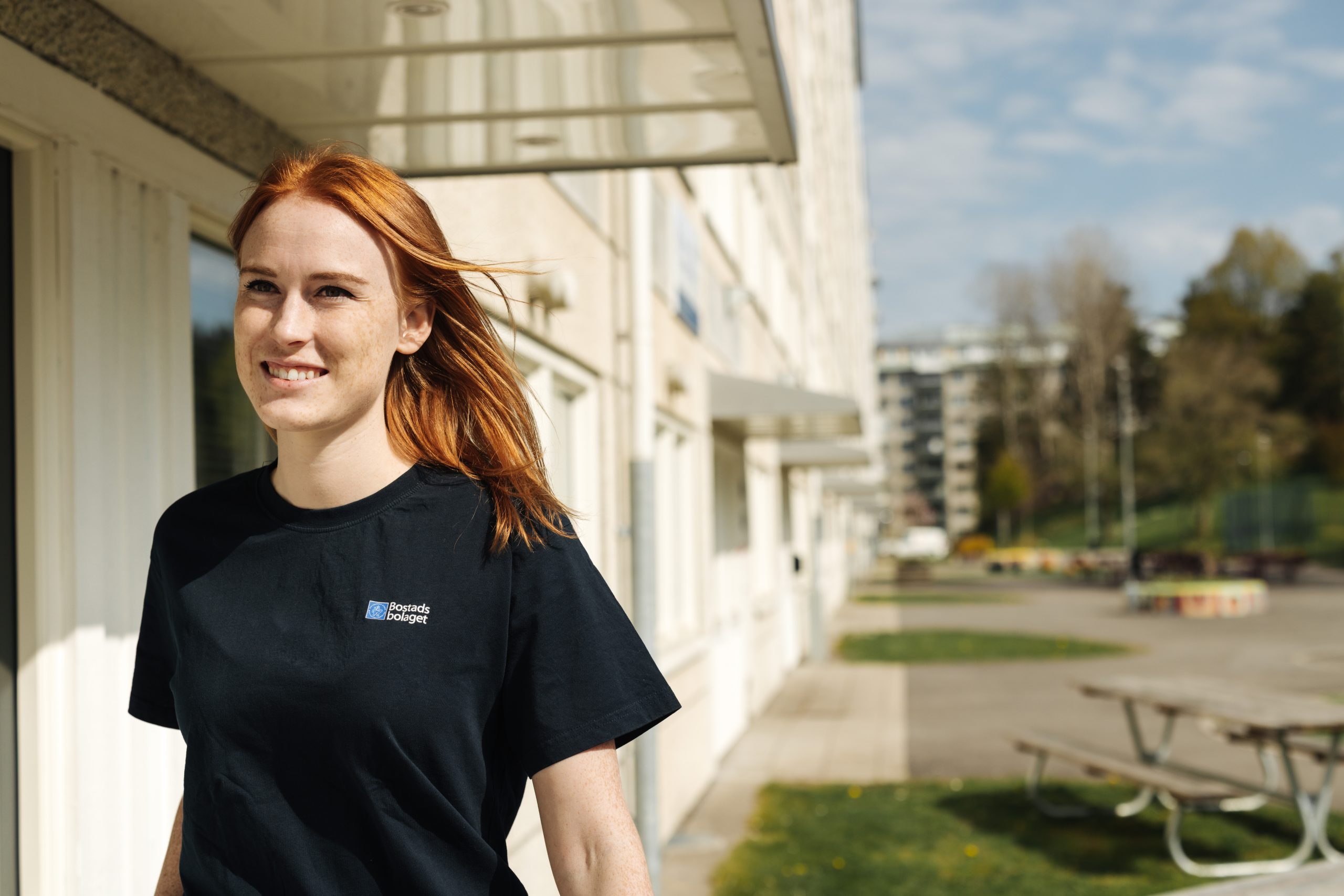 En ung kvinna står framför en byggnad och ler, iklädd Bostadsbolagets arbetskläder.