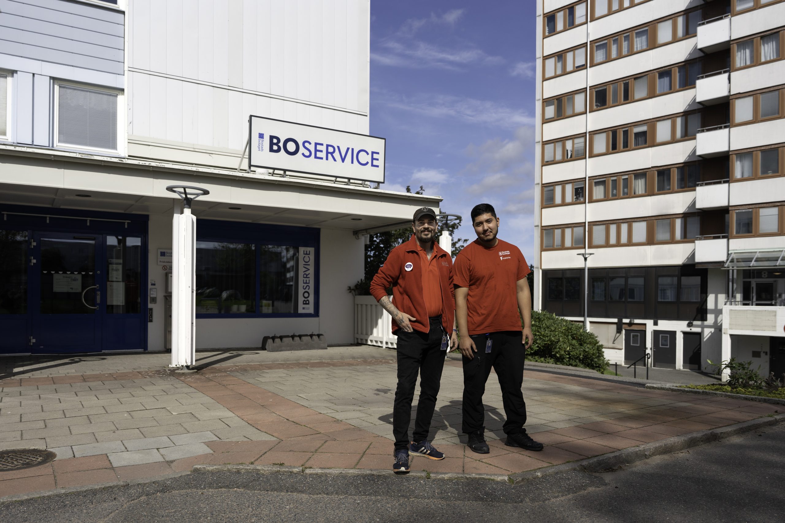 Bostadsbolagets trygghetsvärdar Mio och Masoud iklädda Bostadsbolagets arbetskläder. De står framför ett Boservice och tittar in i kameran och ler.
