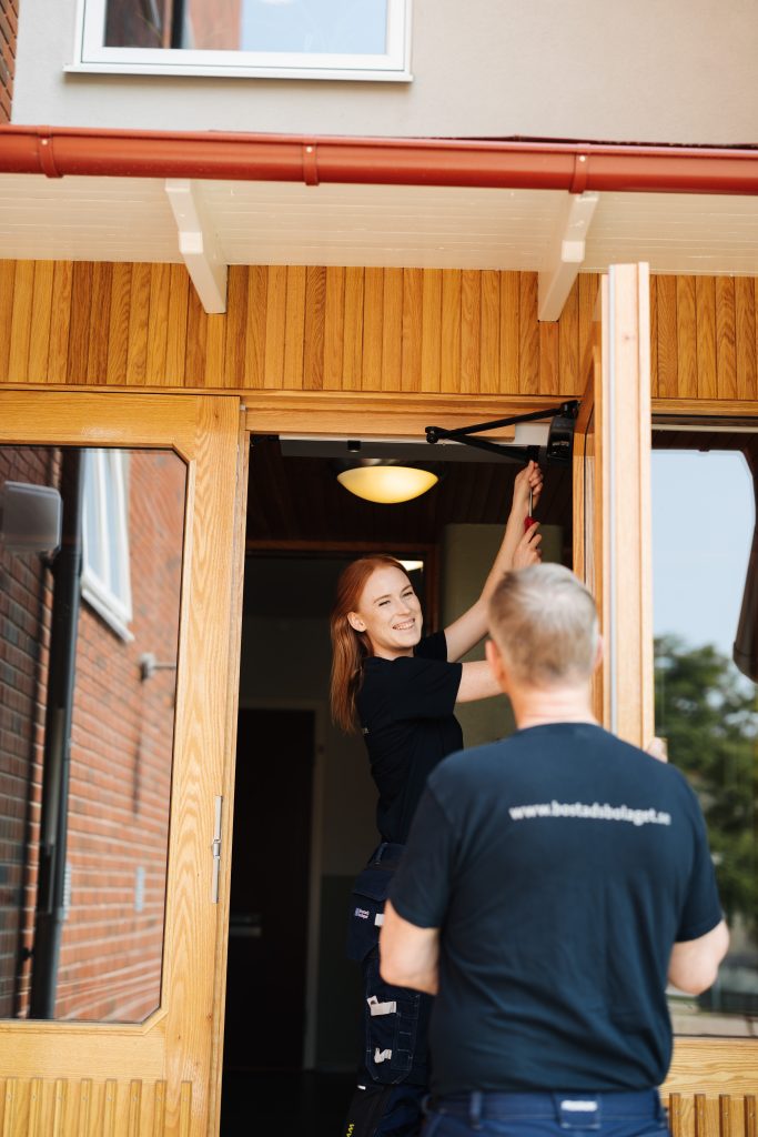 Ung kvinna står i en port till en lägenhetsbyggnad och ler mot sin kollega.