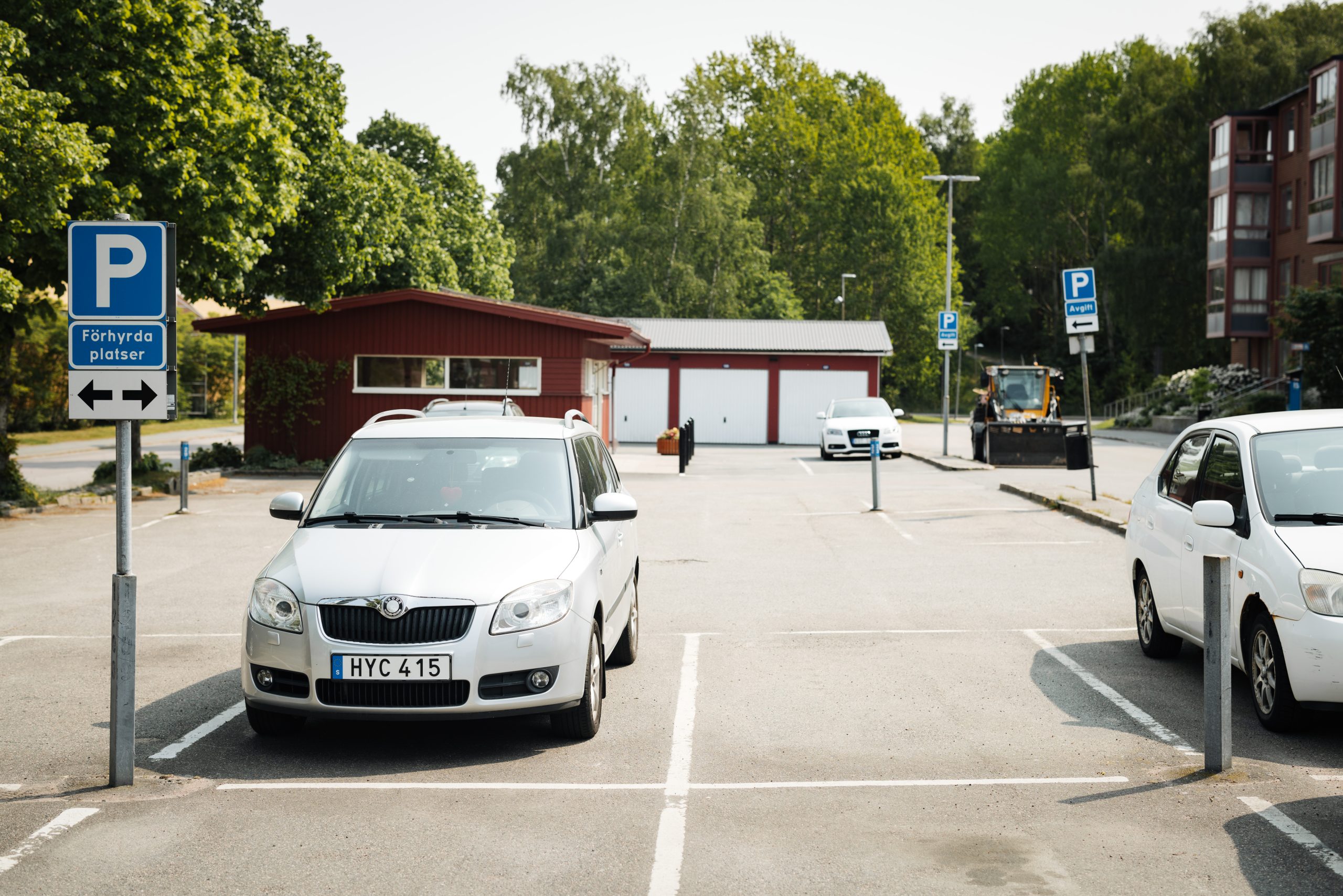 Två vita bilar som står parkerade på en parkeringsplats. I bakgrunden syns en parkeringsskylt, ett miljörum och gröna träd. 