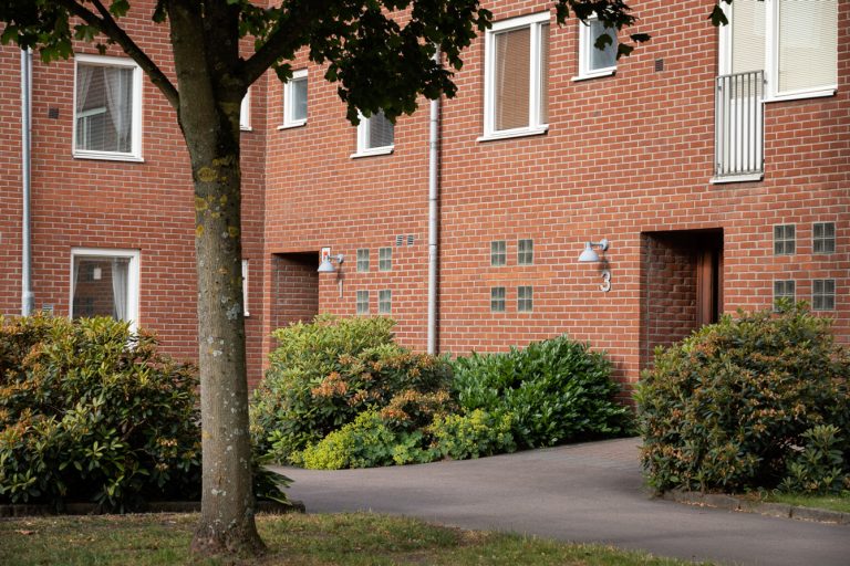 Ett hyreshus i tegel med träd och buskar framför.