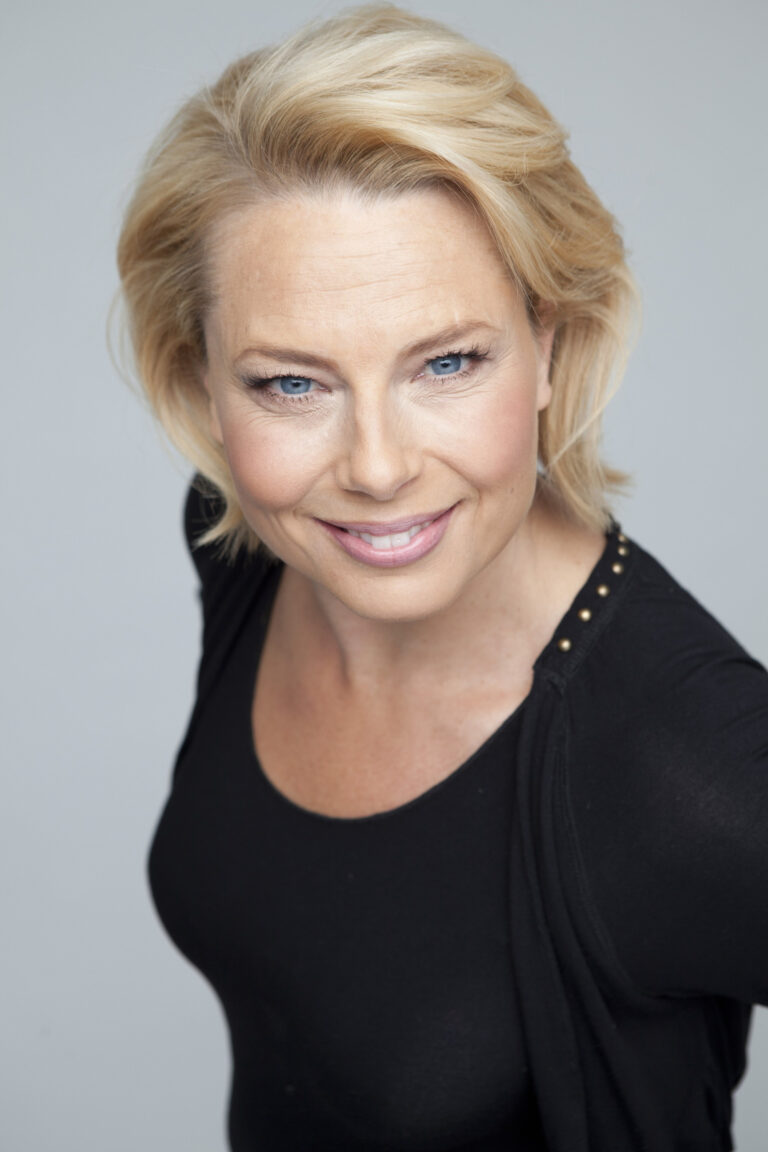 En kvinna med blont hår och blå ögon som kollar in i kameran.