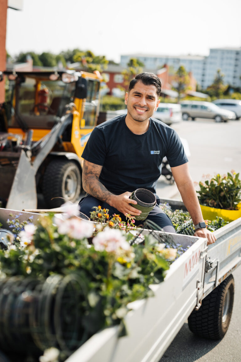 En man med Bostadsbolagets arbetskläder sitter i ett släp och planterar blommor.