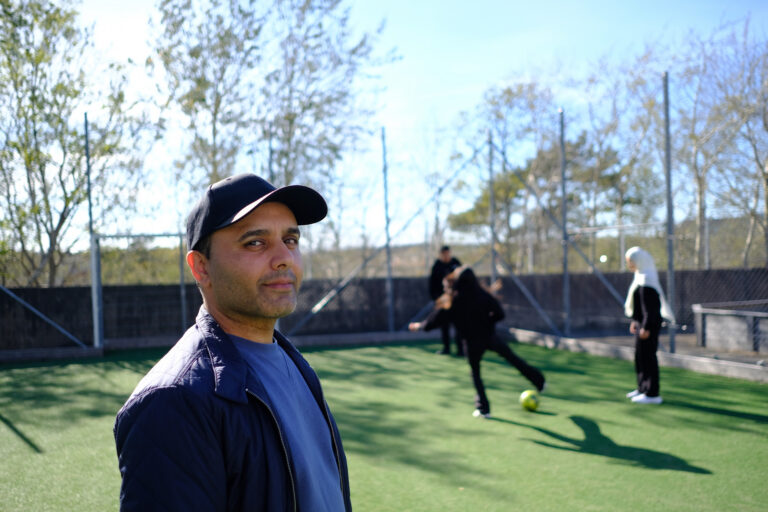 Fotbollstränaren Ibrahim står på en fotbollsplan och kollar in i kameran, iklädd mörkblå kläder och keps. I bakgrunden spelar några ungdomar fotboll.