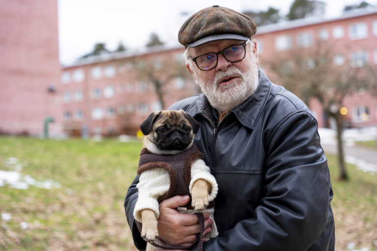 En äldre man i basker håller i en mops. I bakgrunden syns ett par rosa lägenhetshus och en gräsmatta.