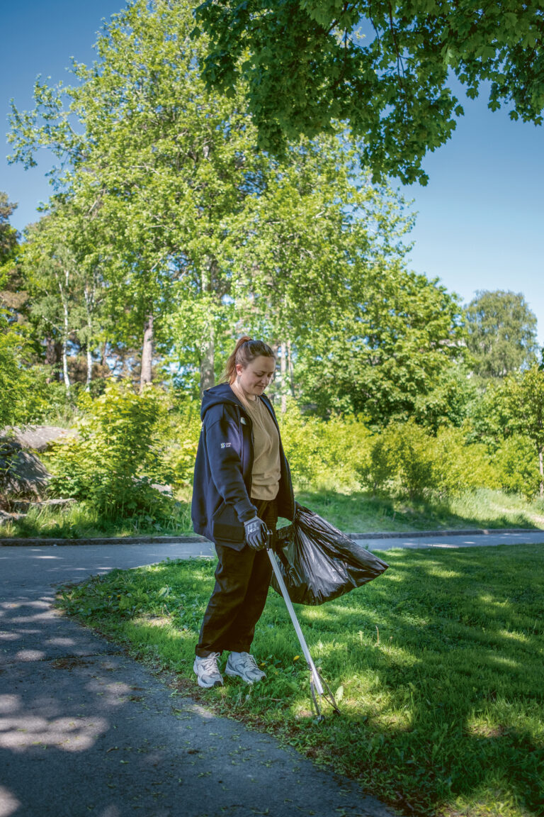 En kvinna i en park med en sopborste och en påse.