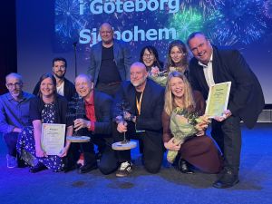 Nio glada människor på en scen med blommor, diplom och pris.