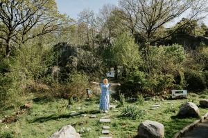 Kvinna i blå klänning står i en trädgård och sträcker ut armarna. 