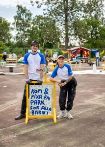 En kille och en tjej står framför en handmålad skylt där det står Kom och fixa en park för alla barn.