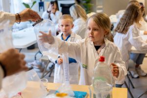 Liten flicka i vit labbrock deltar i ett vetenskapligt experiment. Hon håller upp en plastpåse som blåsts upp. I bakgrunden finns andra barn.