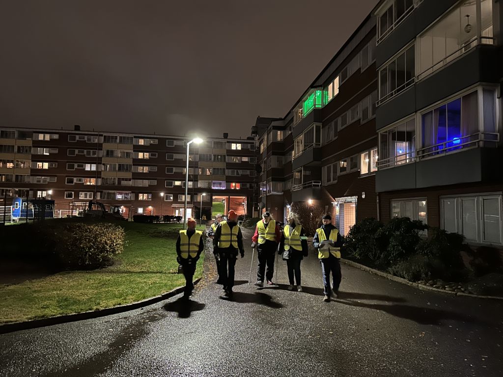Fem personer med gula reflexvästar går i ett mörkt bostadsområde.
