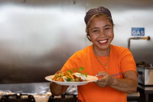 En kvinna ler in i kameran och håller en tallrik mat i handen.