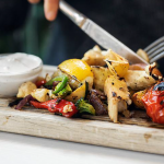 Kött, potatis coh grönsaker ligger på en skärbräda. Någon skär med kniv och gaffel i maten.