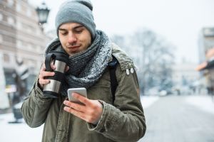 Ung man går utomhus i vintertid med en kaffemugg och tittar i sin telefon.