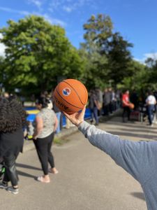 Folk vid basketplanen tittar på en mtach. I bildens fokus syns en basketboll.