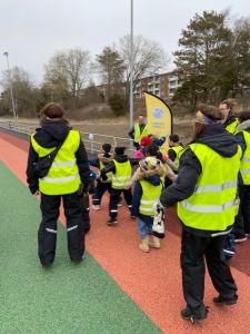 Funktionärer och barn i gula västar står med ryggen mot kameran på en löparbana.