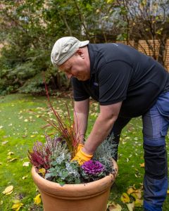 En man i arbetskläder planterar blommor i en stor kruka utomhus.