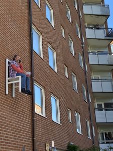 en karaktär i utställningen x-times people chair pratar i telefon och äter jordgubbar på en stol som är fastskruvad i fasaden. Två personer fotar honom från sina balkonger.